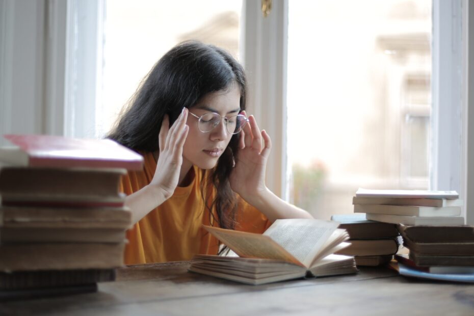 Une jeune femme assise à son bureau entre des piles de livres se tient les tempes, les yeux fermés, pour se concentrer