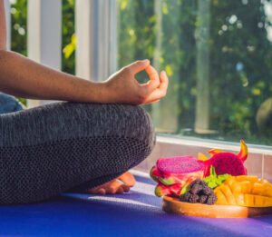 Une femme en posture du lotus est assise à côté d'un plateau de fruits exotiques, mangues, fruits du dragon...