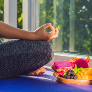Une femme en posture du lotus est assise à côté d'un plateau de fruits exotiques, mangues, fruits du dragon...